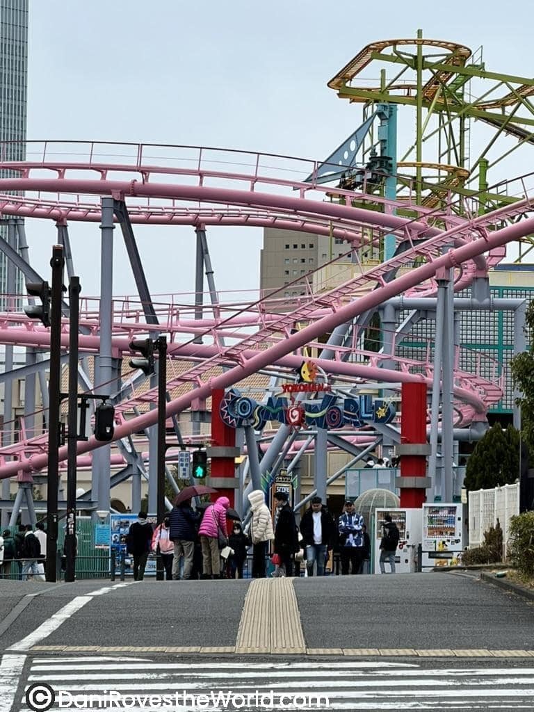 : The entrance to Yokohama’s Cosmo World amusement park, showcasing vibrant pink roller coaster tracks and the colorful “Cosmo World” sign, with visitors bundled up on a chilly day.