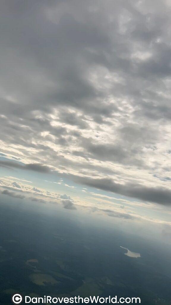 An aerial view of thick, layered clouds seen from an airplane window. The clouds obscure much of the view, creating a moody and overcast sky.
