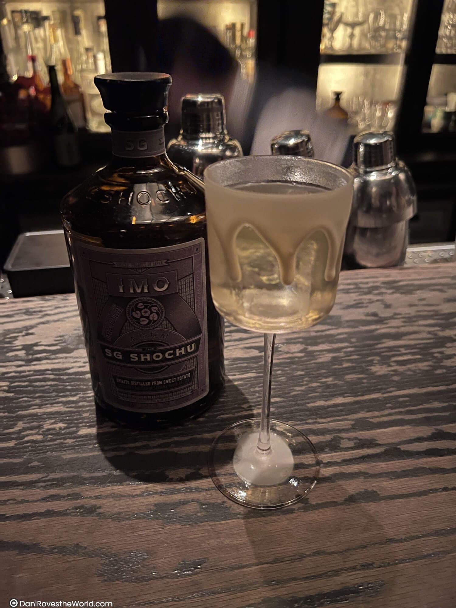 A bottle of SG Shochu, a spirit distilled from sweet potato, displayed on a rustic wooden bar. Next to the bottle is a delicate cocktail glass with an elegant design, featuring what appears to be a frothy, creamy layer near the rim. The dimly lit bar setting adds a warm and inviting ambiance, with cocktail shakers and shelves of bottles visible in the background