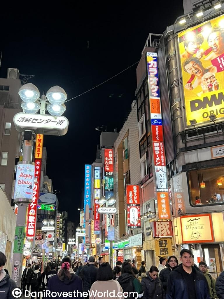 A vibrant, crowded street in Shibuya at night, lined with bright neon signs advertising restaurants, shops, and entertainment venues. The street is bustling with people under the glow of urban lights.