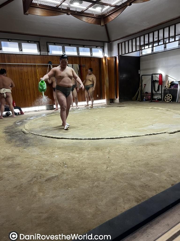 A sumo wrestler walks across the clay ring, watering the surface using a green watering can, while others rest and watch in the background.
