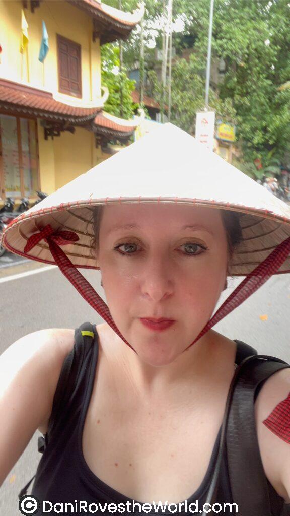 A person wearing a traditional conical hat, with a black tank top and a backpack, poses on a street lined with lush greenery and East Asian architecture in the background.