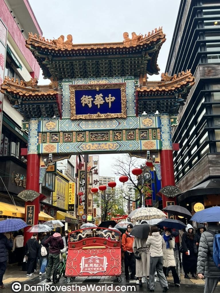 The ornate entrance gate of Yokohama Chinatown, featuring intricate blue, green, and gold details, red columns, and a crowded street beneath umbrellas.