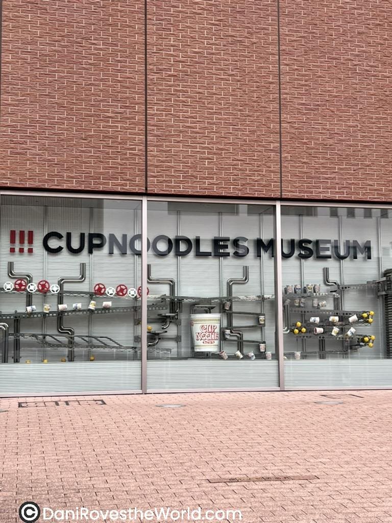 The front window of the Cup Noodles Museum with intricate piping designs displaying cup noodles as part of the exhibit, set against a red brick exterior.