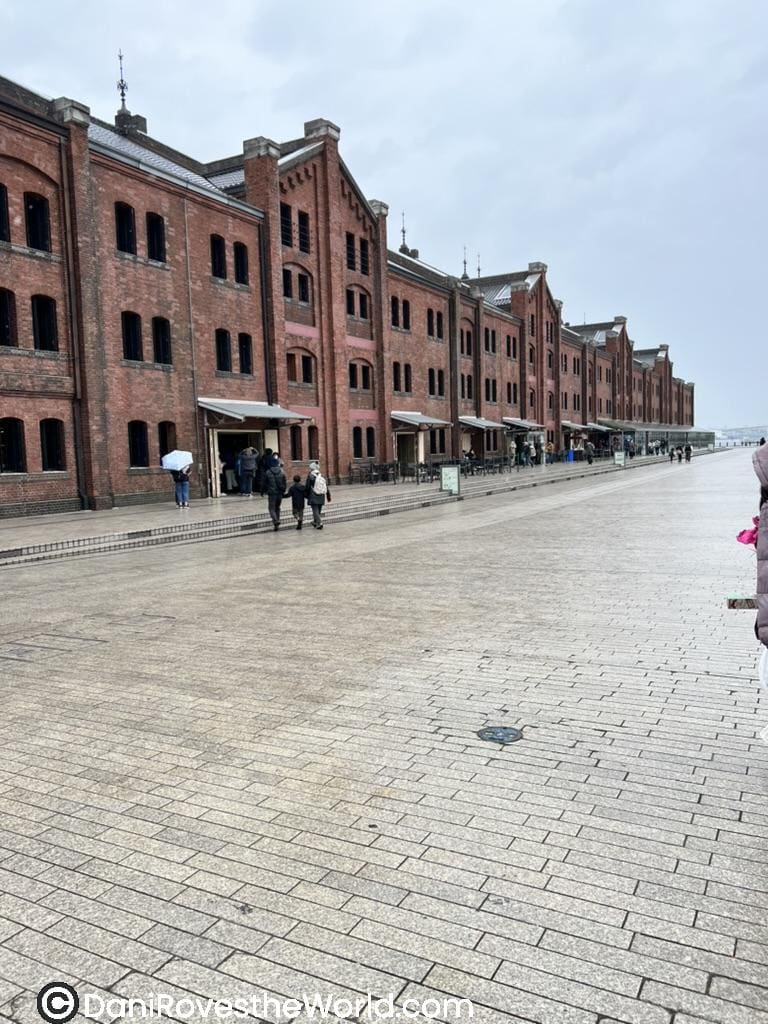 The historic red brick warehouses in Yokohama line a wide, wet pavement, with visitors strolling and umbrellas in use under an overcast sky.