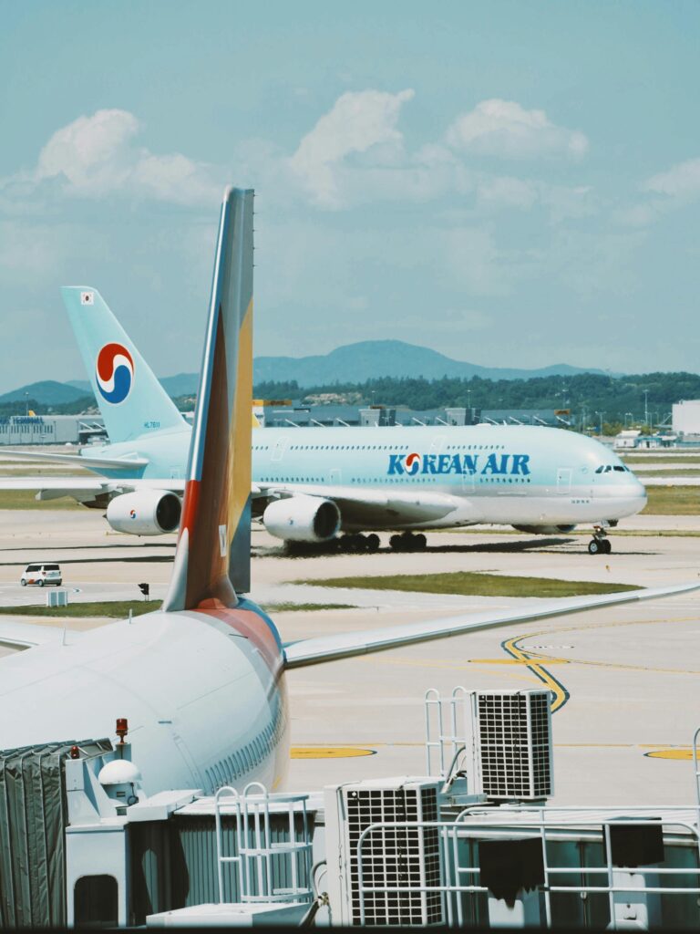 A Korean Air plane on a sunny airport runway with a smaller aircraft tail in the foreground. The scene captures the busy airport backdrop with a mountain range in the distance under clear skies.
