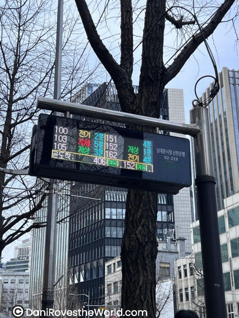 Digital sign at a Seoul bus stop displaying real-time bus arrival information in Korean, surrounded by high-rise buildings—highlighting efficient public transport during January in Seoul.