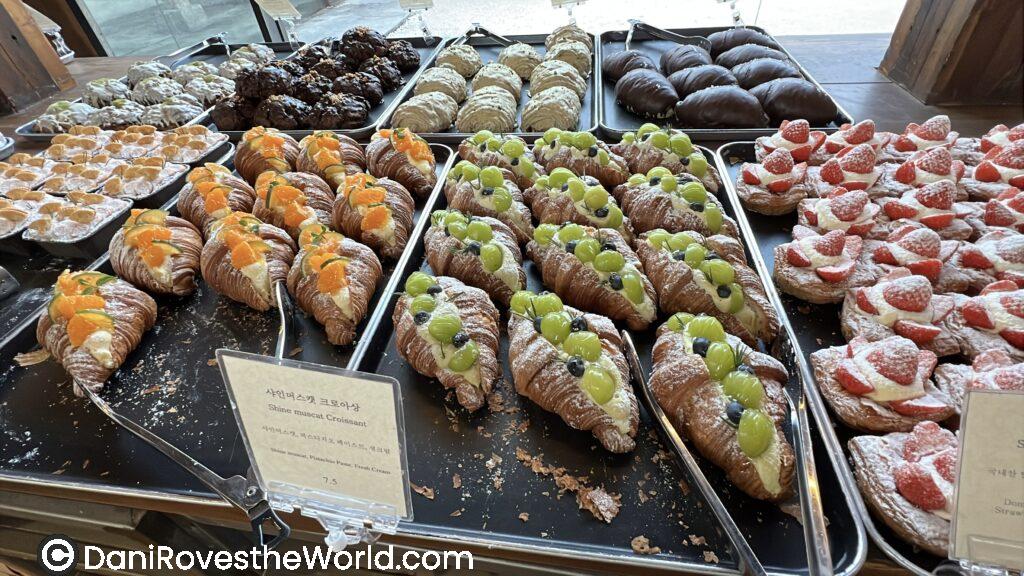 Display trays in a Seoul coffee shop are filled with colorful pastries, including croissants topped with green grapes, orange slices, and powdered sugar, showcasing a vibrant selection of fruit-topped desserts.