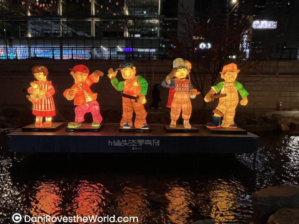 Illuminated lantern figures of people in winter attire line a streamside platform during a nighttime Lantern Festival in Seoul, a vibrant highlight of January in Seoul.