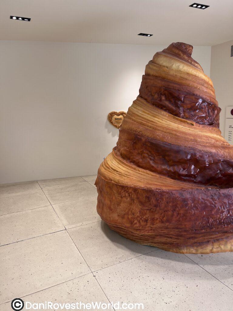 Inside a trendy coffee shop in Seoul, a quirky white mannequin sculpture kneels while holding croissant-themed boxes and signage, positioned near a giant croissant installation in a warm-toned minimalist interior with tiled floors and natural sunlight casting rainbows.