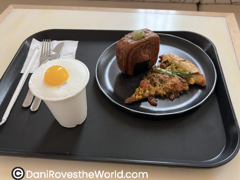 A black tray at a coffee shop in Seoul holds a unique selection: a white cup topped with fluffy foam and a yolk-like yellow topping, a cube-shaped pastry with a green garnish, and a slice of savory pastry with corn, green beans, and herbs.
