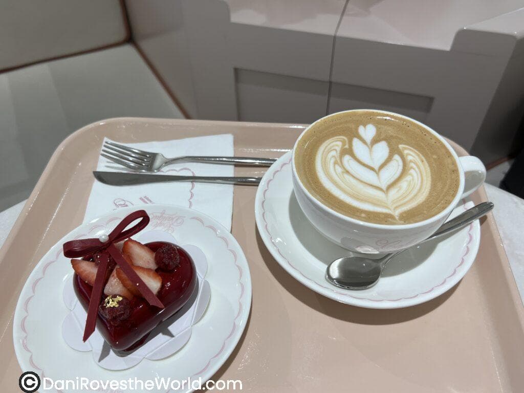 On a soft pink tray at a chic coffee shop in Seoul, a heart-shaped mousse dessert decorated with strawberries and ribbon is paired with a latte adorned with delicate leaf-shaped latte art.