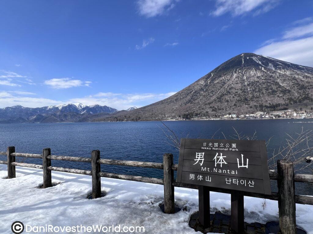 View of Mt. Nantai rising behind a frozen lakeshore in Nikko National Park, with a wooden sign marking the scenic viewpoint—perfect for nature lovers taking a winter day trip from Tokyo.