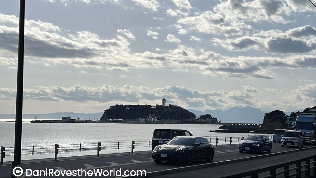 A scenic view of Enoshima Island seen from the coastal road, with cars driving along the water’s edge and a lighthouse rising above the island’s silhouette—an easy seaside getaway from Tokyo.