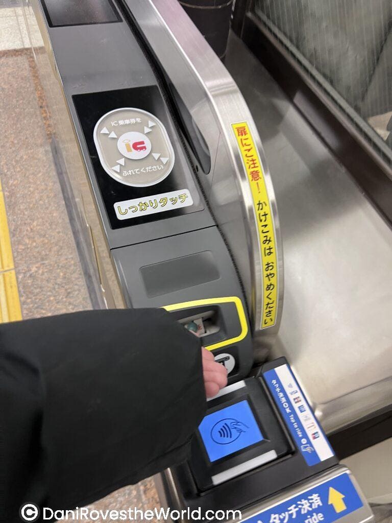 A person taps a contactless transit card at a Tokyo subway gate reader, preparing to enter the train system. The ticket gate displays Japanese instructions and a blue reader screen for IC card payment, a common method for navigating public transportation in Tokyo.