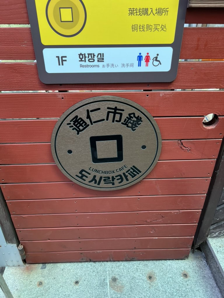 A wooden wall at Tongin Market featuring a round sign shaped like a traditional Korean brass coin, labeled “Lunchbox Cafe” in English and Korean. Above it, a yellow restroom sign in multiple languages indicates facilities for men, women, and people with disabilities on the first floor.