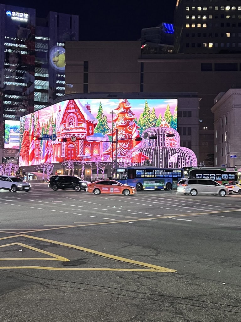 Large digital billboard in Seoul at night displays a kaleidoscopic light show with star and jewel-like patterns in blue, white, and red hues. Below, pink-lit trees and a dome-shaped light installation glow near passing buses and cars, creating a festive urban scene.