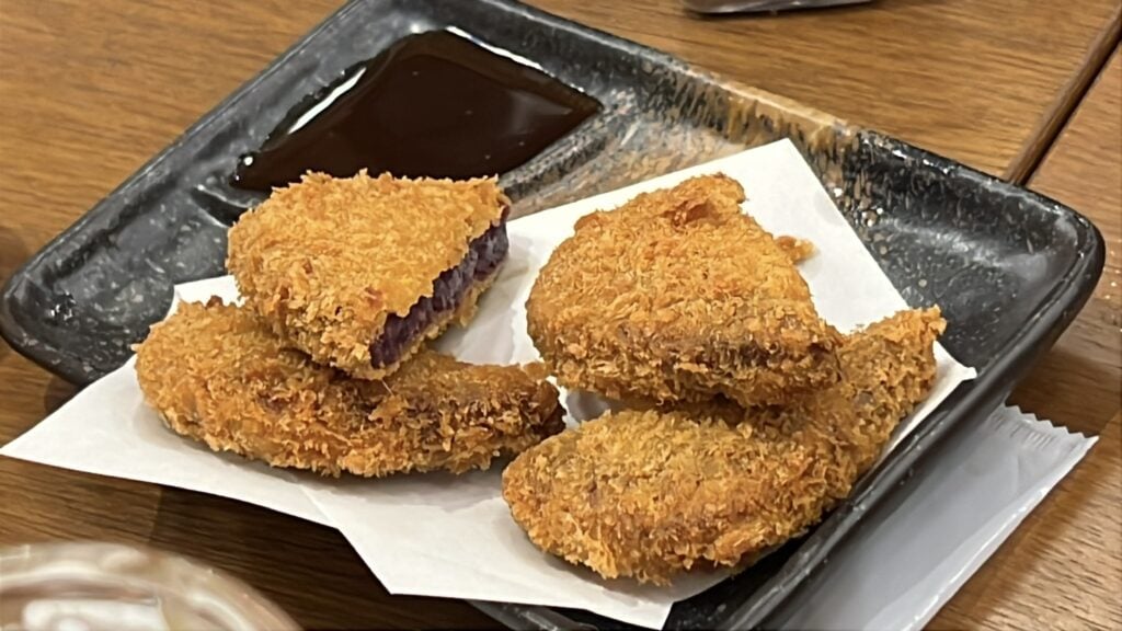 Crispy golden croquettes filled with vibrant purple sweet potato, served on a black plate with tonkatsu sauce during a Tokyo izakaya tour.