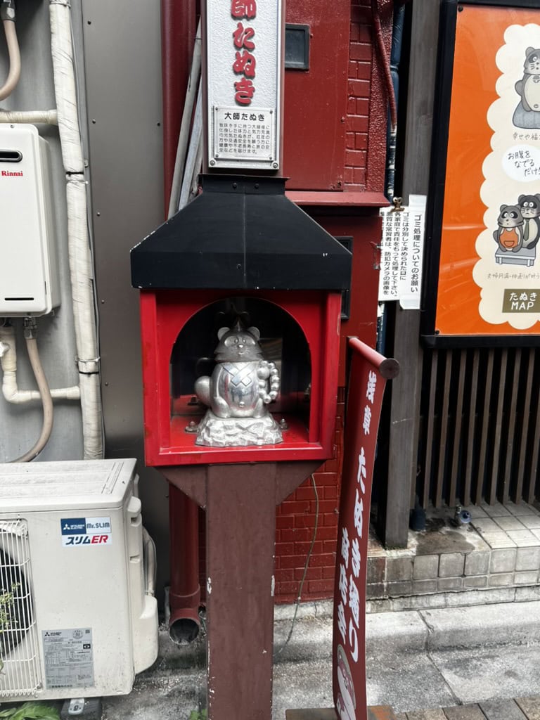 Small tanuki statue enshrined in a red and black case on a quiet Asakusa street, with surrounding signs in Japanese describing its cultural meaning. A quirky local sight you might stumble upon while exploring Asakusa.