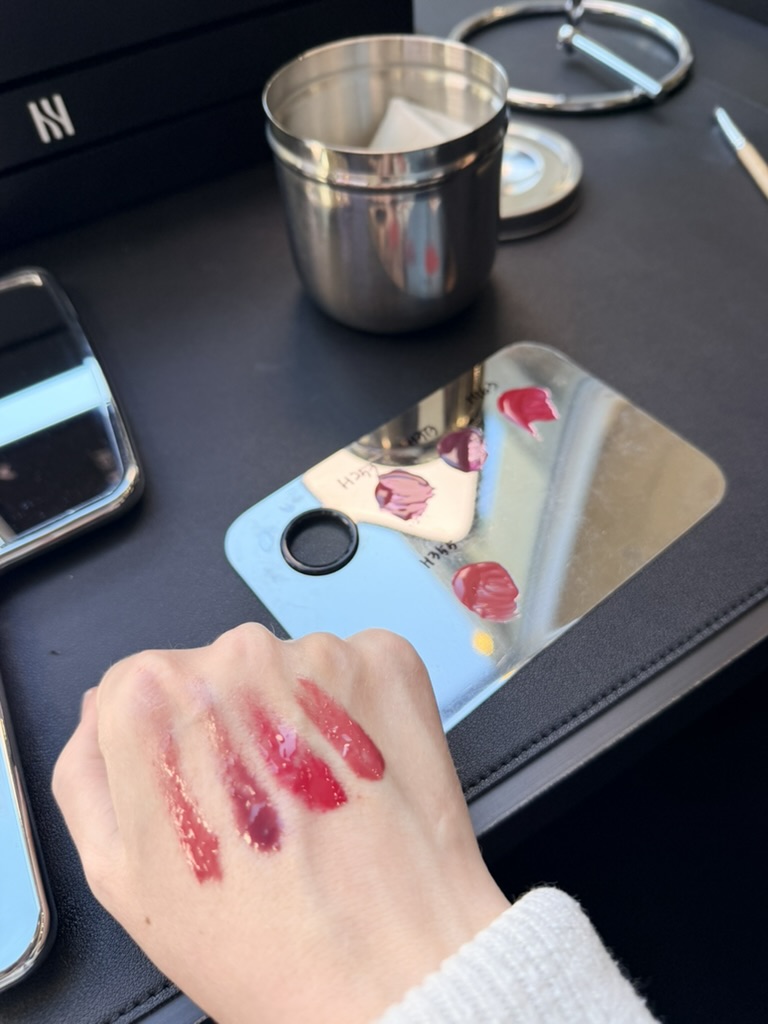Hand swatched with multiple glossy lip colors in red and berry shades next to a makeup palette and tools.