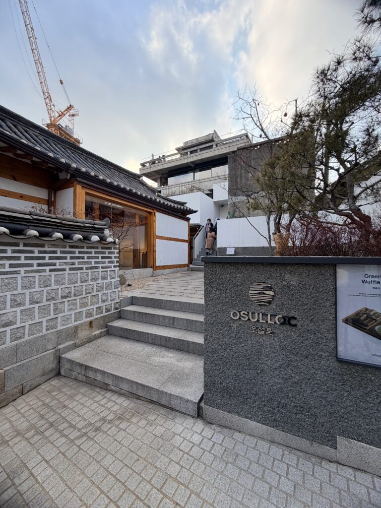 Entrance to Osulloc Tea House in Seoul with traditional hanok architecture, stone steps, and modern buildings in the background.